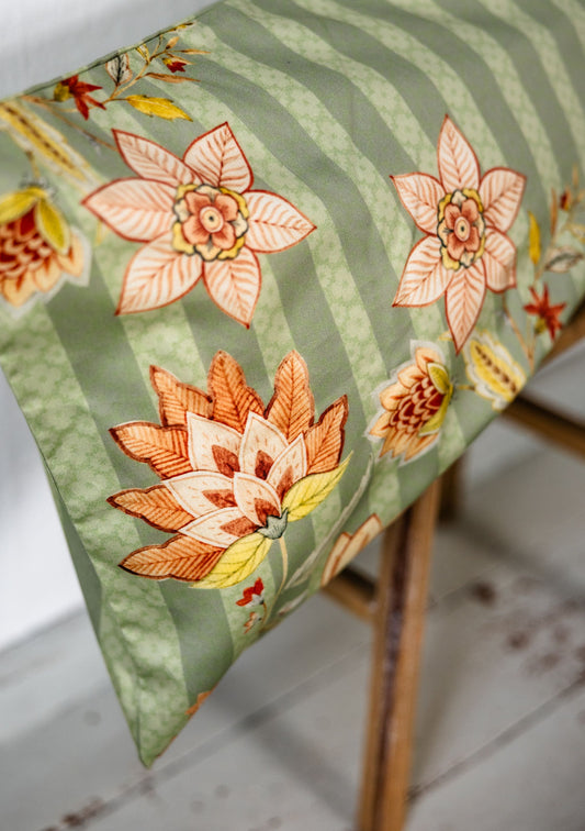 Detailaufnahme eines grünen Bettkissens mit großem floralen Muster in Orange, Gelb und Rosé, auf einem Holzschemel.