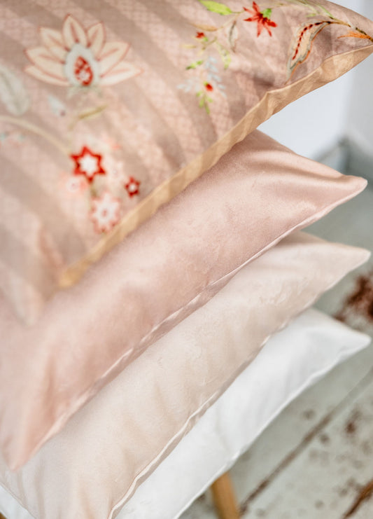 Detailaufnahme eines Kissenstapels mit floraler Vorderseite oben und darunter edle Premium-Samt-Kissen in Rosé-Beige und Cremeweiß – weiche Haptik und elegante Farbabstufungen.