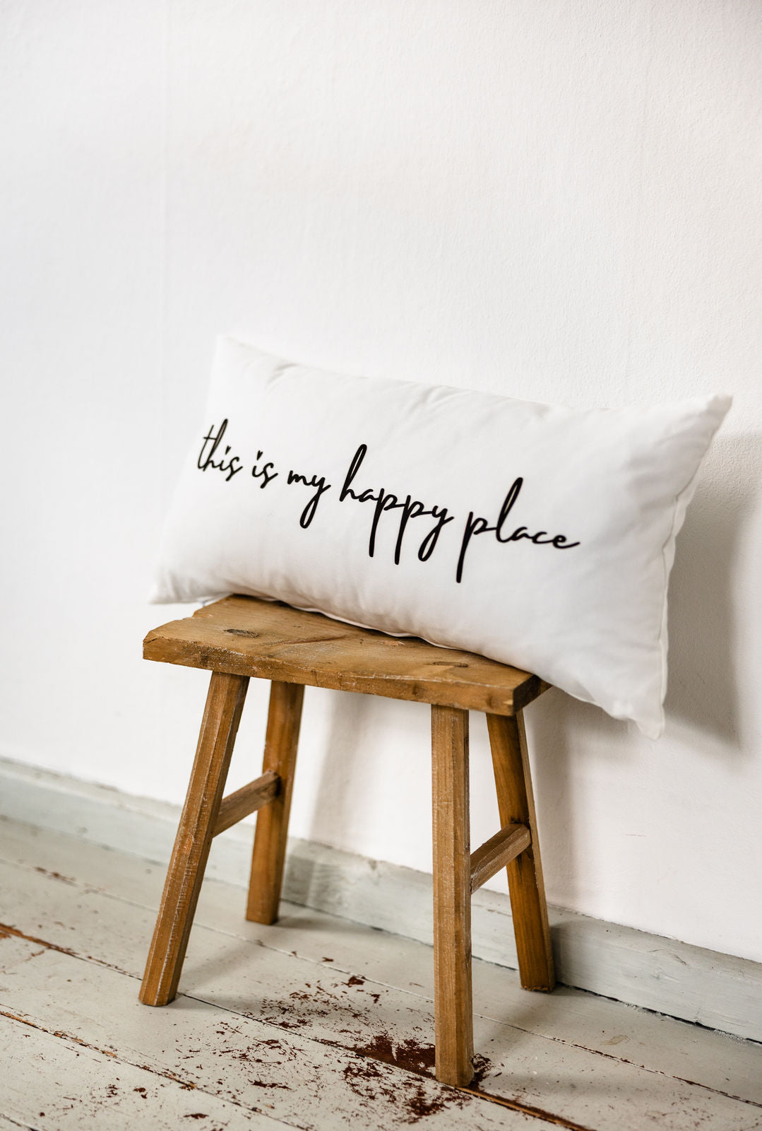 Weißes Dekokissen mit schwarzem Schriftzug „this is my happy place“ im Format 30x60 cm, locker auf einem rustikalen Holzstuhl vor heller Wand arrangiert.