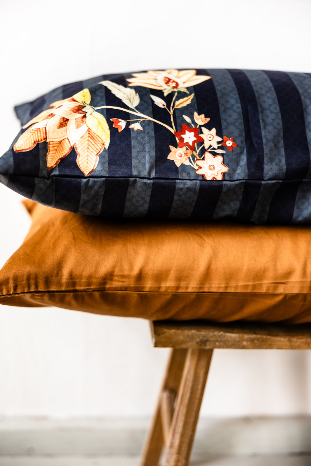 Detailansicht zweier gestapelter rechteckiger Bettkissen im Format 40x60 cm auf rustikalem Holzstuhl vor weißer Wand: oben dunkelblau mit Streifen- und Blumenmuster, unten unifarben in Cognac.