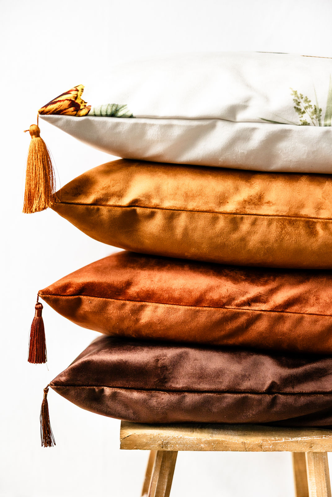 Vier gestapelte Premium-Samt-Dekokissen mit Quasten in den Farben Weiß mit Blumenmuster, Cognac, Kupfer und Schokoladenbraun, seitlich fotografiert auf einem rustikalen Holzstuhl.