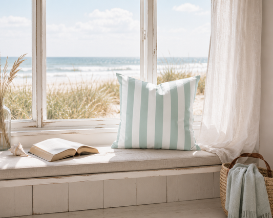 Dekokissen mit Aqua-Blau-Weißen Blockstreifen auf einer Fensterbank mit Blick auf Strand und Meer. Einrichtungsidee für Ferienwohnung, Ferienhaus und Küstenstil an der Ostsee.