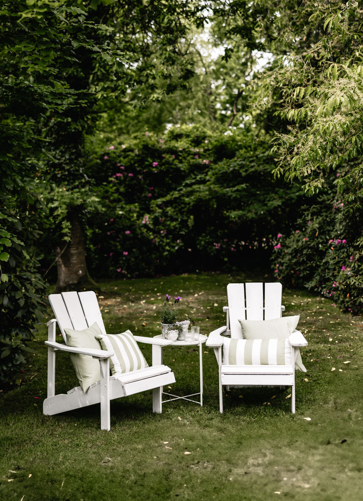 Zwei weiße Gartenstühle mit grün-weißen Wendekissen auf einer Wiese – gemütliche Outdoor-Ecke unter Bäumen
