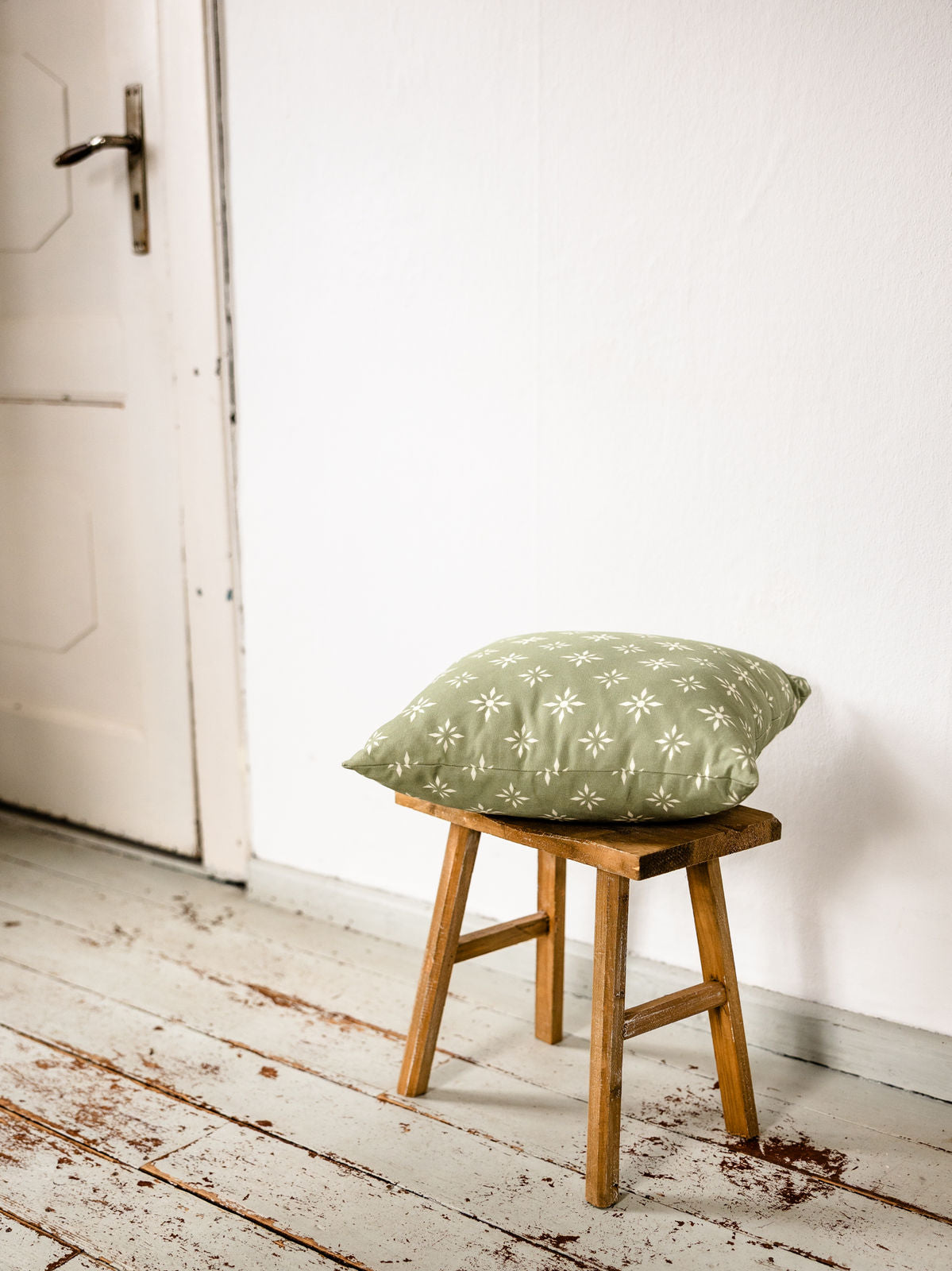 Salbeigrünes Kissen mit weißem skandinavischen Muster, schräg auf einem Holz-Hocker platziert – aufgenommen in einer hellen Raumecke mit weißer Wand und alter Tür.