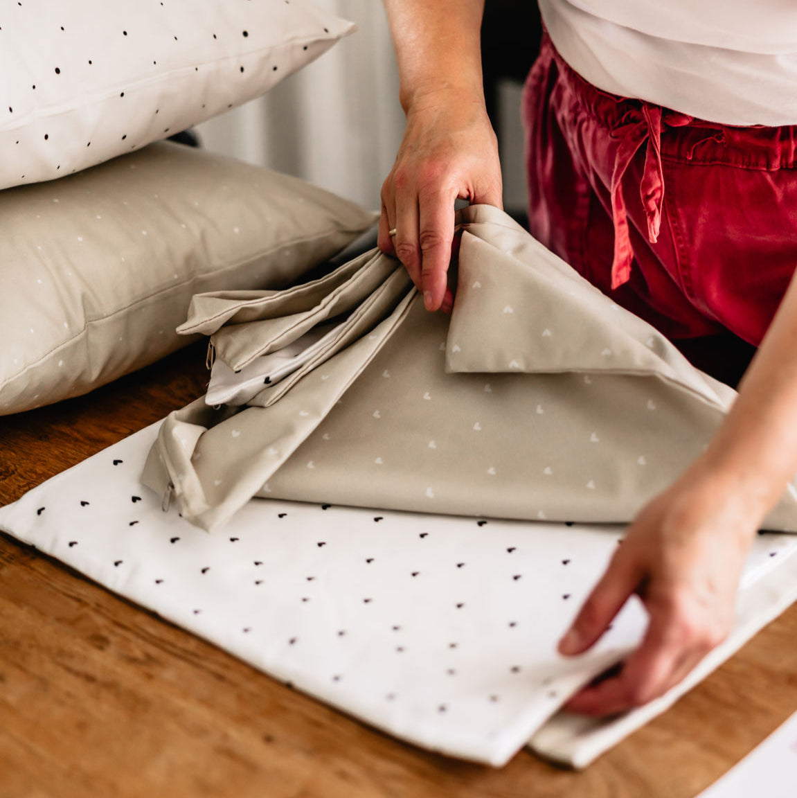 Eine Person faltet Bettwäsche in Beige und Weiß mit feinem Herzmuster – aus weichem Baumwoll-Perkal, daneben passende Kissenhüllen im selben Design auf einem Holztisch.