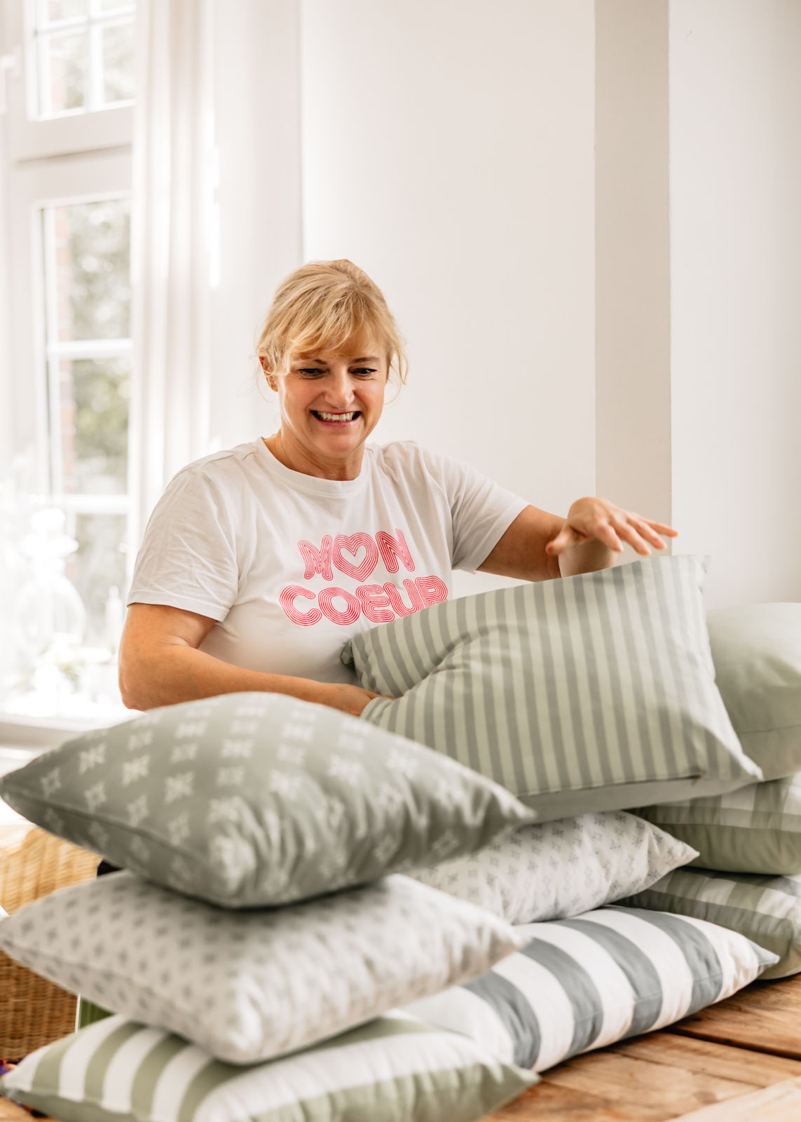 Shopinhaberin Marion Christensen von „Christensen – Scandinavian Home Design“ in hellem Raum mit Fenster, beim Drapieren eines grün gestreiften Kissens auf einem hohen Kissenstapel aus verschiedenen grünen Designs