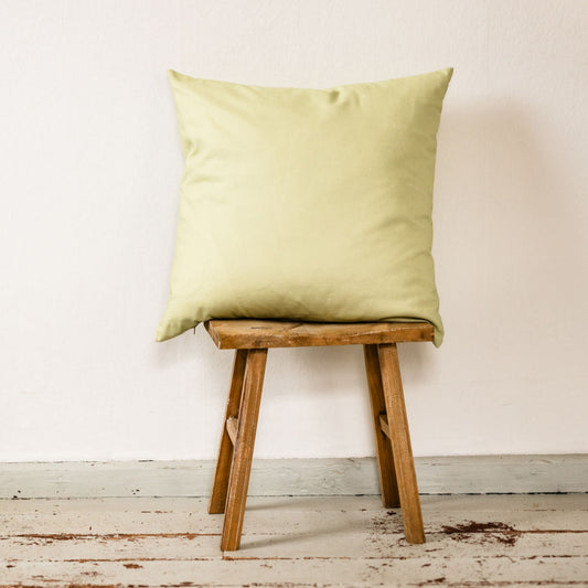 Einfarbiges Dekokissen aus sanftgrüner Baumwolle in 50 × 50 cm, schlichtes Design – ideal zum Kombinieren mit gemusterten Kissen, auf rustikalem Hocker vor heller Wand