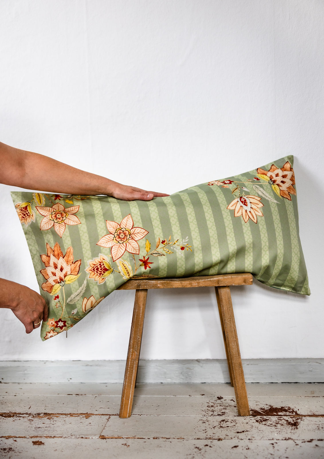  Grünes Bettkissen mit floralen Mustern wird von zwei Händen auf einem Holzschemel positioniert, vor schlichter weißer Wand.