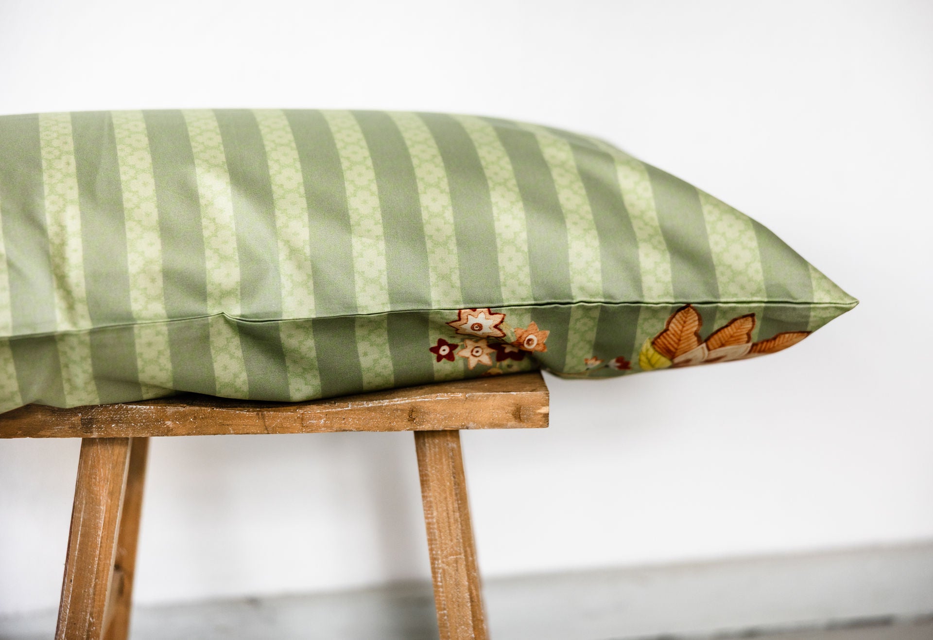  Rückseite eines grünen Bettkissens aus Baumwollperkal mit Ton-in-Ton gehaltenen Blockstreifen, auf einem Holzschemel platziert.