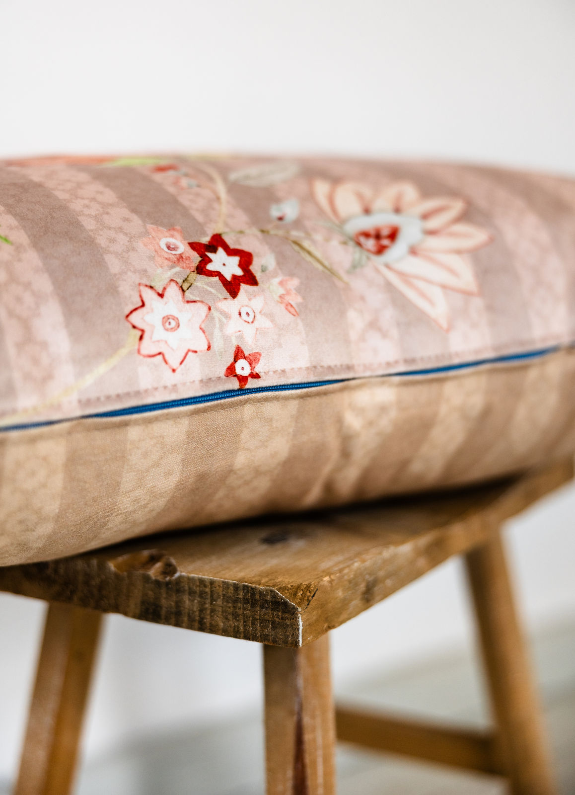 Detailansicht eines rosé-beigen Blumenkissens mit floraler Stickerei und türkisfarbenem Reißverschluss, auf rustikalem Holzschemel platziert
