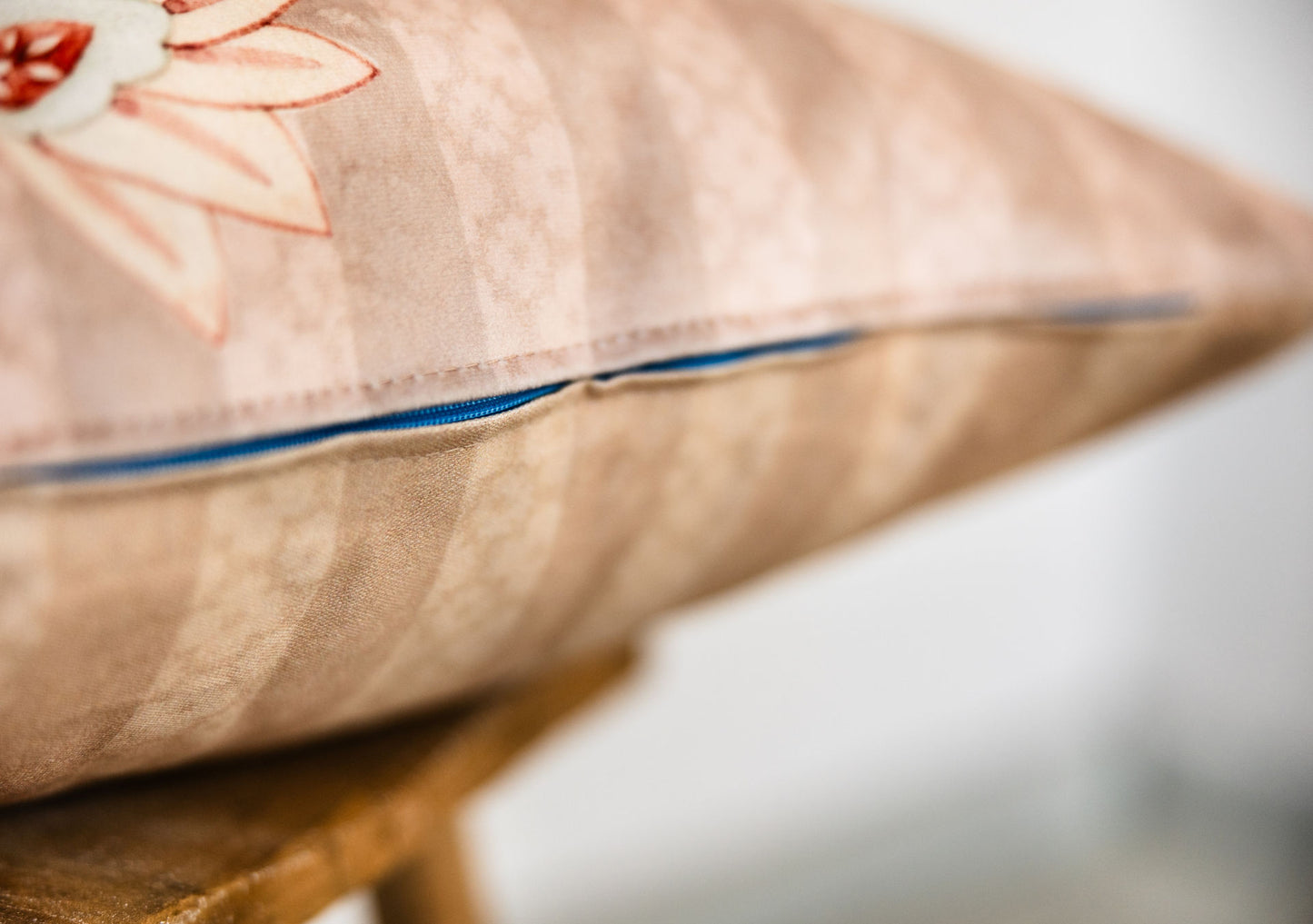 Detailaufnahme des türkisblauen Reißverschlusses an rosé-beigem Zierkissen mit floralem Druck und dezentem Streifenmuster auf Holzschemel