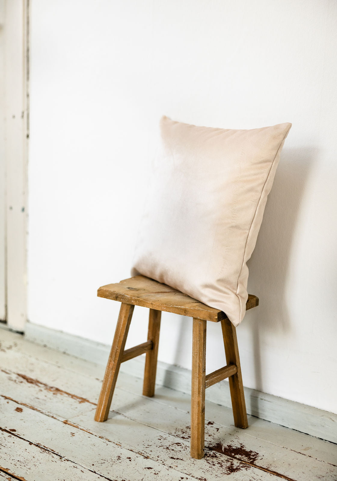 Kissen in Soft White aus schimmerndem Premium-Samt, seitlich auf rustikalem Holzschemel vor weißer Wand fotografiert