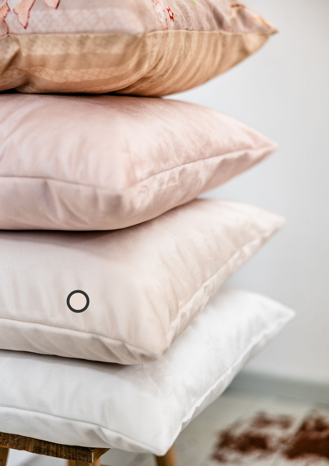 Stapel aus vier Premium-Samtkissen in Rose-Beige, Creme- und Weißtönen – auf einem Hocker vor weißer Wand arrangiert.