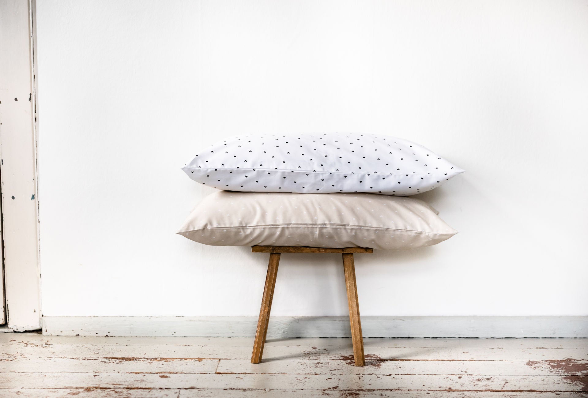 Zwei 80x40 cm große Bettkissen auf einem Holzstuhl: oben ein weißes Kissen mit schwarzen Herzchen, darunter ein beiges Kissen mit weißen Herzchen – schlichtes, liebevolles Design aus Baumwoll-Perkal.