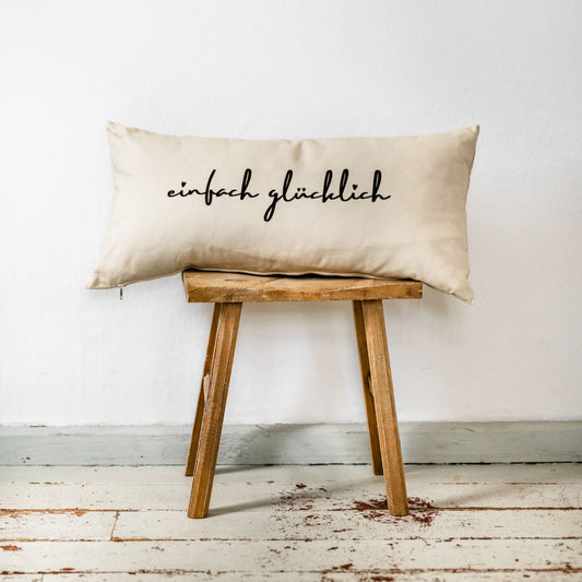 Beiges Dekokissen im Format 30x60 cm mit handschriftlichem Schriftzug „einfach glücklich“ in Schwarz, aus Baumwollstoff, dekorativ auf einem rustikalen Holzstuhl platziert.