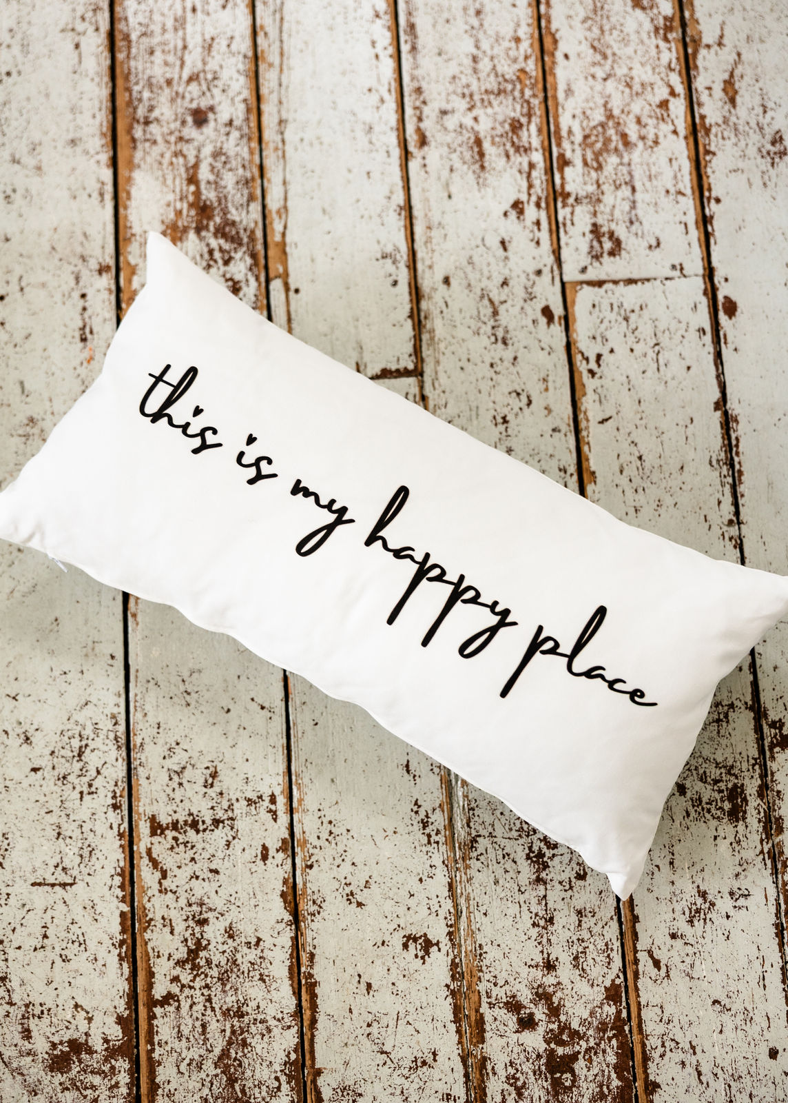 Weißes Dekokissen im Format 30x60 cm mit schwarzem Schriftzug „this is my happy place“, schräg auf rustikalem Holzboden arrangiert, von oben fotografiert.