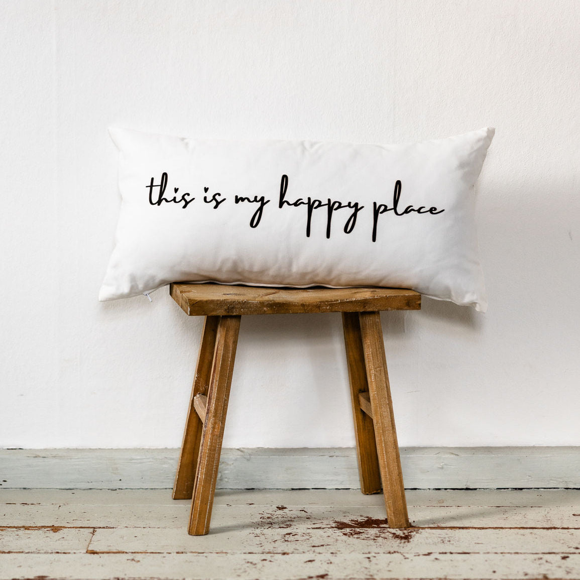 Weißes Dekokissen im Format 30x60 cm mit schwarzem handschriftlichem Schriftzug „this is my happy place“, dekorativ auf einem Holzstuhl vor weißer Wand platziert.