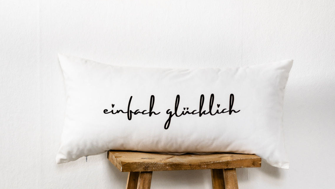 Weißes Dekokissen im Format 30x60 cm mit schwarzem Schriftzug „einfach glücklich“, aus Baumwolle, dekorativ auf einem rustikalen Holzstuhl vor heller Wand platziert.