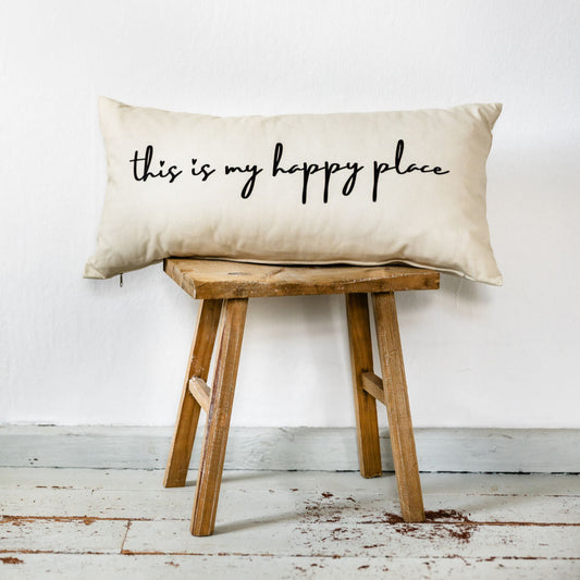 Beiges Dekokissen im Format 30x60 cm mit schwarzem handschriftlichem Schriftzug „this is my happy place“, dekorativ auf einem Holzstuhl vor heller Wand platziert.