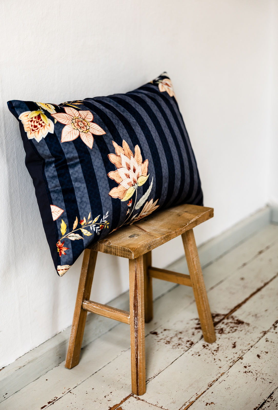  Dekokissen im Format 40×60 cm mit dunkelblauen Streifen und floralen Blütenmustern in Rosé- und Beigetönen auf der Vorderseite, Rückseite unifarben dunkelblau, angelehnt auf rustikalem Holzschemel vor weißer Wand.