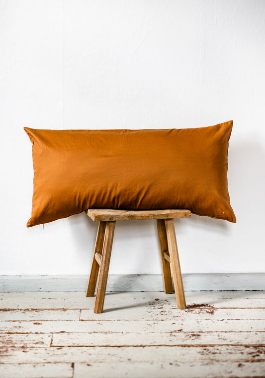 Rechteckiges Bettkissen im Format 40x60 cm in einfarbigem Cognac-Ton aus Baumwoll-Perkal, liegend auf rustikalem Holzstuhl vor weißer Wand.