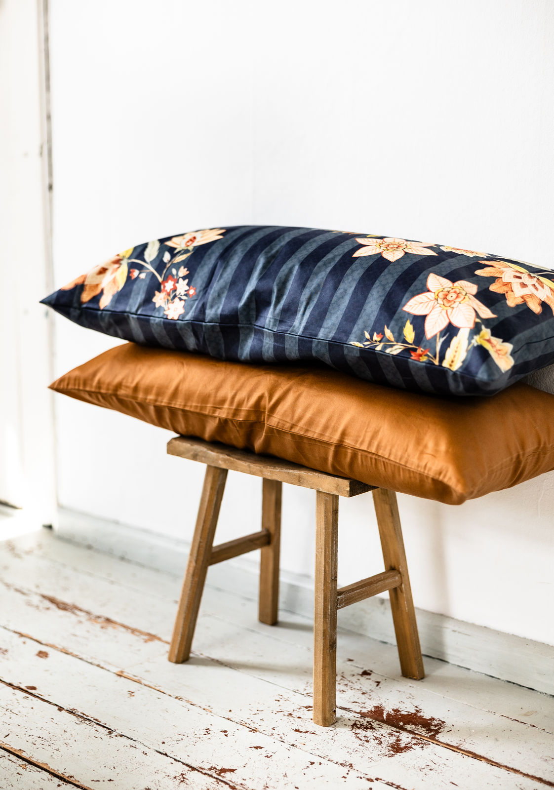 Zwei rechteckige Bettkissen im Format 40x60 cm, seitlich auf einem rustikalen Holzstuhl vor weißer Wand gestapelt: oben ein dunkelblaues Kissen mit Streifen- und Blumenmuster, unten ein unifarbenes Kissen in Cognac-Ton.