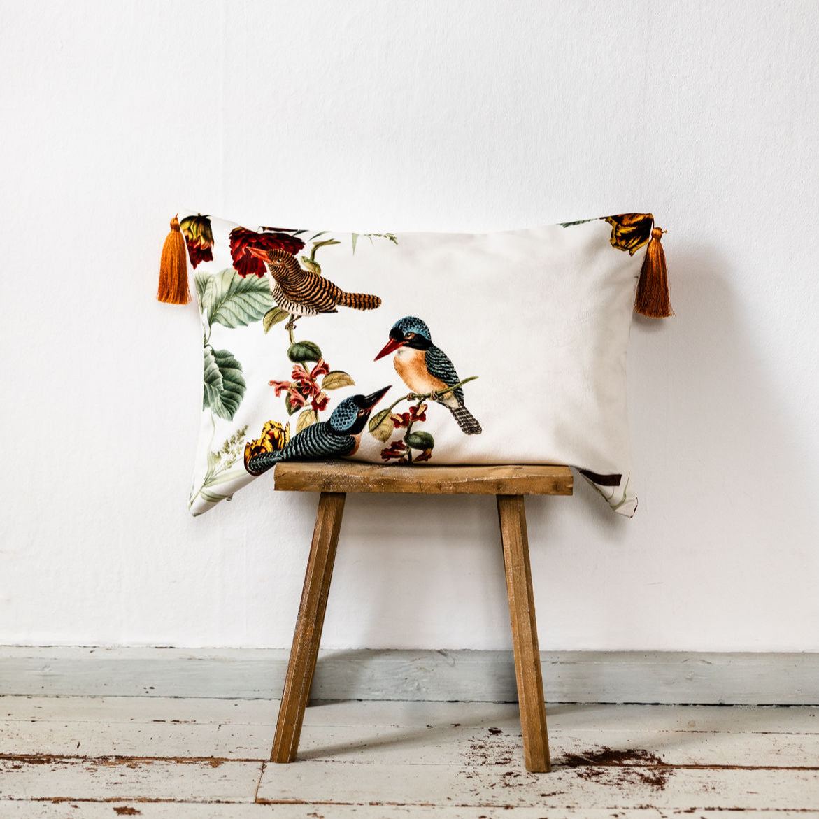 Dekokissen aus Premium-Samt in 40x60 cm mit Vogel- und Blumenmotiv in Weiß und Mehrfarbig, verziert mit goldfarbenen Quasten an den Ecken, platziert auf einem rustikalen Holzstuhl vor weißer Wand.