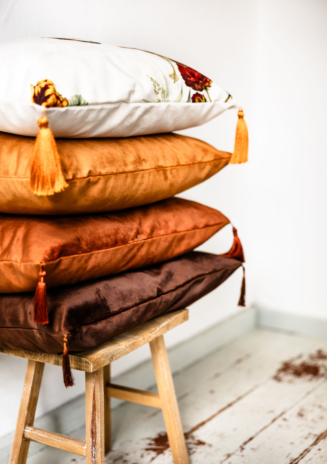 Gestapelte Premium-Samt-Dekokissen mit Quasten in den Farben Weiß mit Blumenmuster, Cognac, Kupfer und Schokoladenbraun, dekorativ auf einem rustikalen Holzstuhl vor weißer Wand platziert.