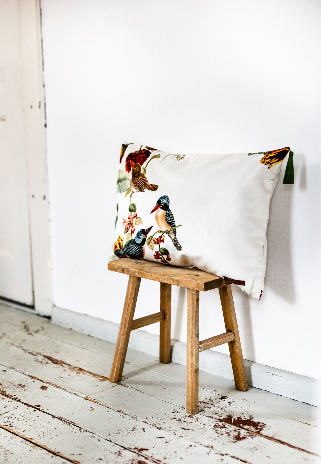 Cremefarbenes Samtkissen mit farbenfrohem Vogel- und Blumenmotiv sowie grünen Quasten an den Ecken, dekorativ auf einem kleinen Holzschemel vor weißer Wand platziert.