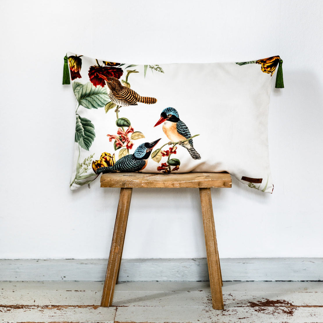 Dekoratives cremefarbenes Samtkissen mit farbigem Vogel- und Blumenprint, verziert mit grünen Quasten an den Ecken, auf einem kleinen Holzschemel vor weißer Wand platziert.