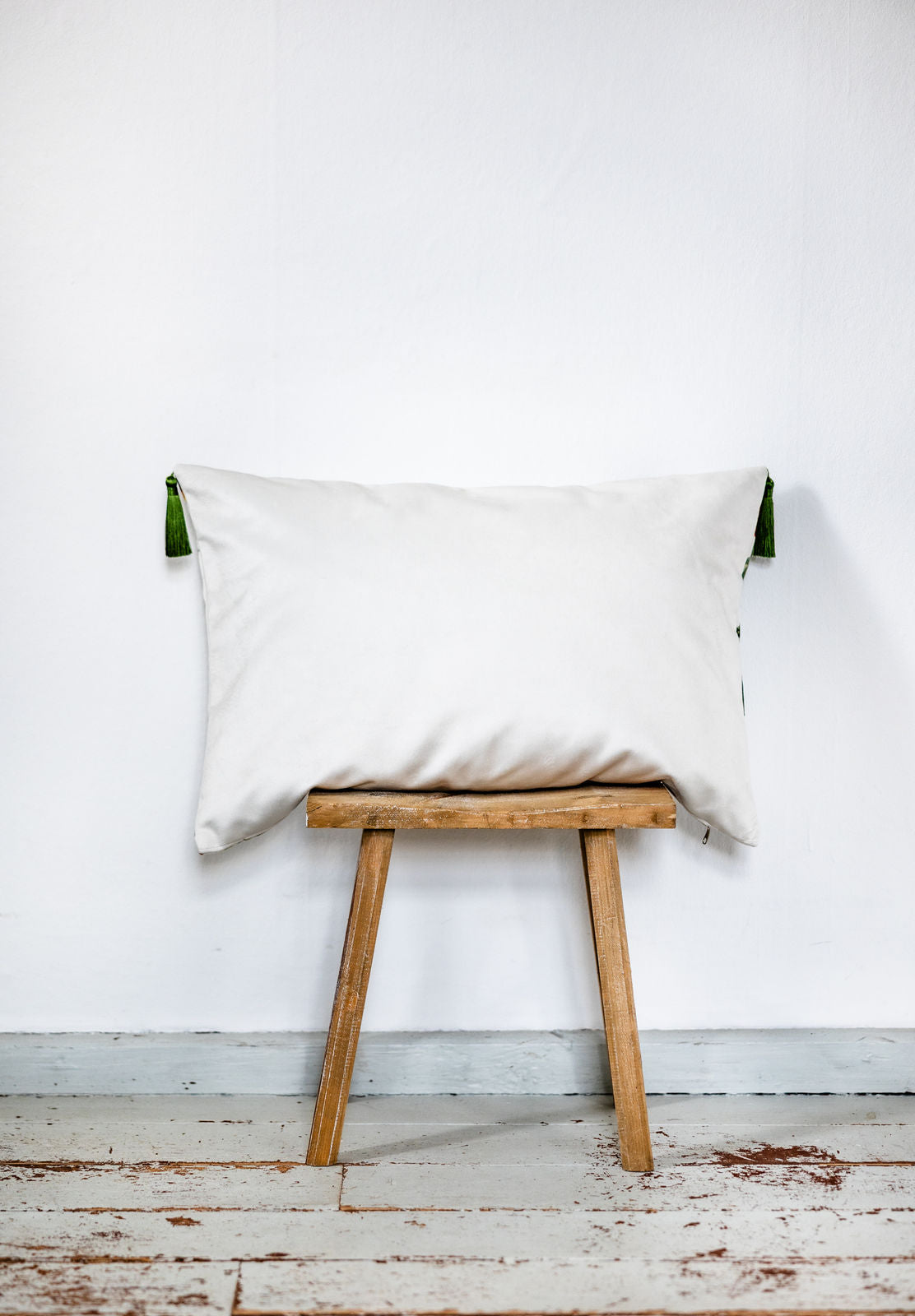 Rückseite eines cremefarbenen Samtkissens mit grünen Quasten an den Ecken, schlicht und elegant auf einem kleinen Holzschemel vor weißer Wand präsentiert.
