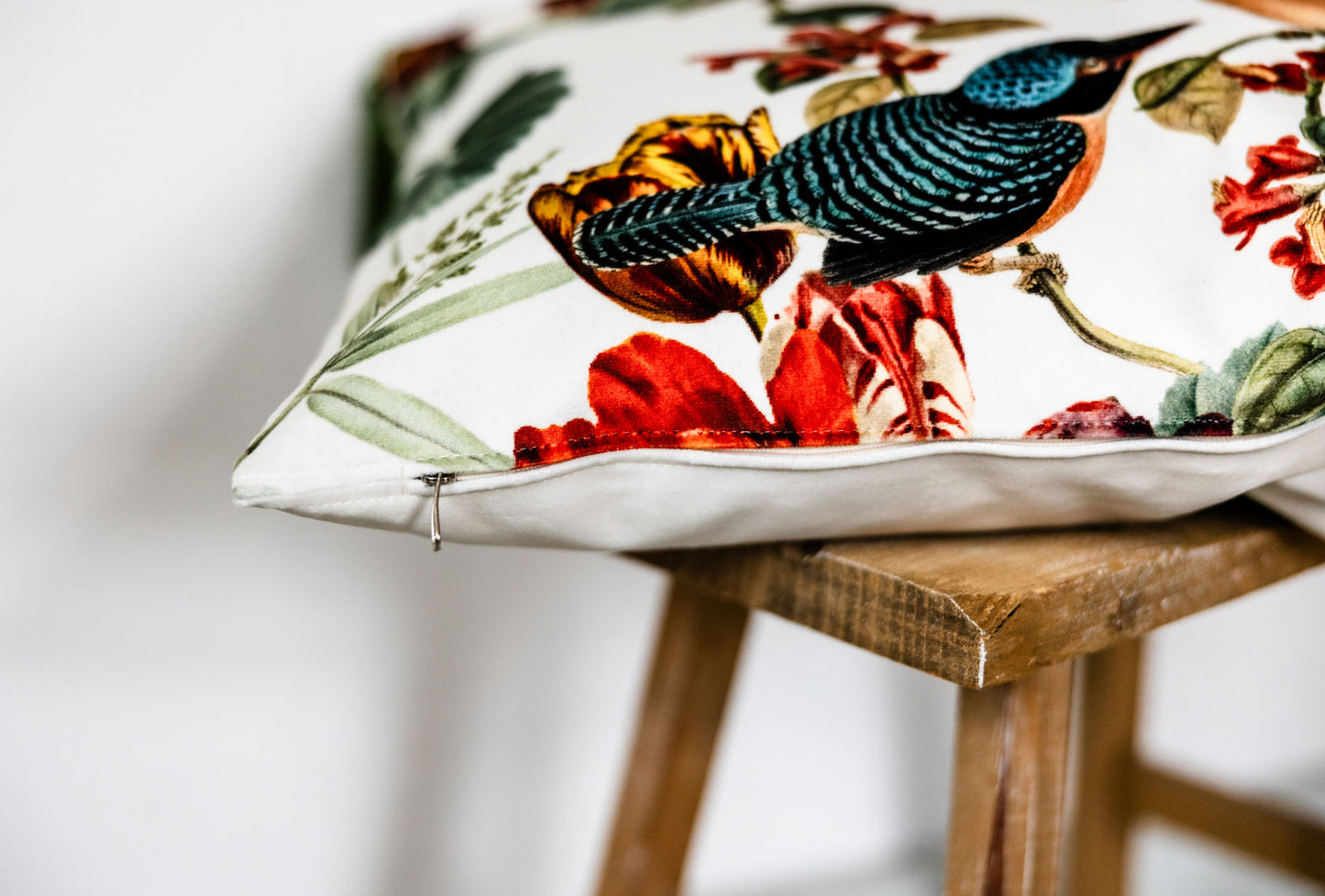 Detailaufnahme eines cremefarbenen Samtkissens mit farbenfrohem Vogel- und Blumenmotiv, sichtbar mit Reißverschluss an der Seite, liegend auf einem kleinen Holzschemel.