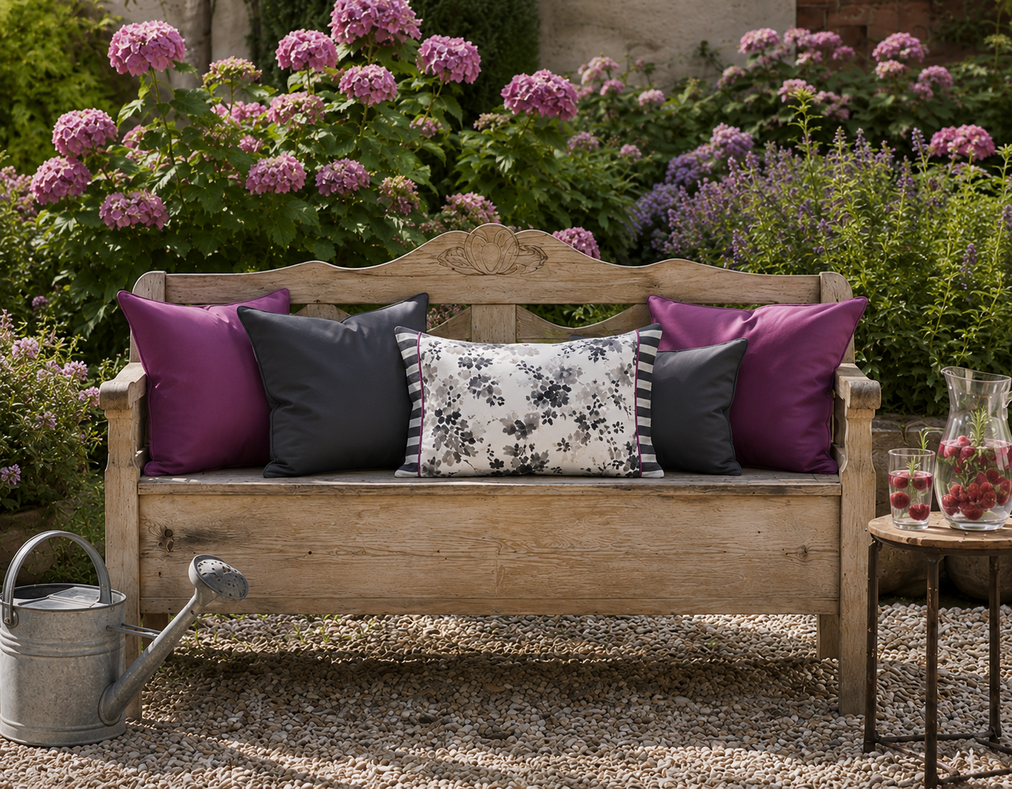 Dekokissen-Kombination in Aubergine, Anthrazit und Weiß auf einer rustikalen Gartenbank. Florales Kissen mit grauem Blütenmuster vor blühenden rosa Hortensien im romantischen Landhausgarten.