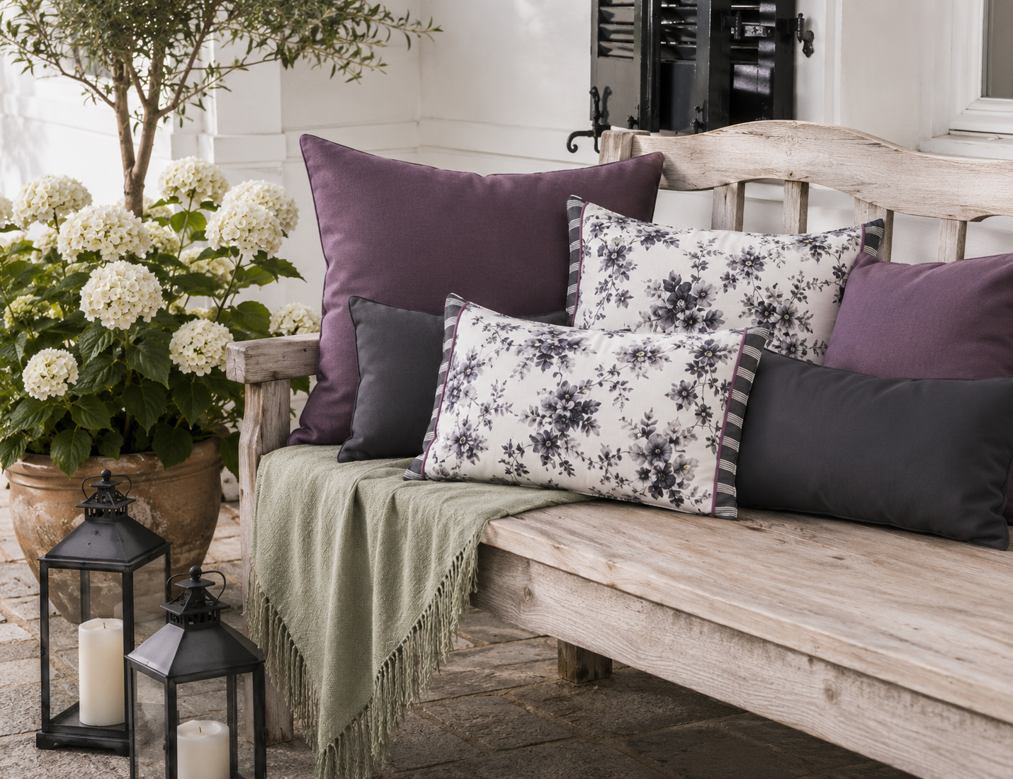 Dekokissen-Kombination in Aubergine, Anthrazit und Weiß auf einer rustikalen Gartenbank. Florale Kissen mit grauem Blütenmuster schaffen eine elegante Atmosphäre im Landhausstil auf Terrasse oder im Garten.