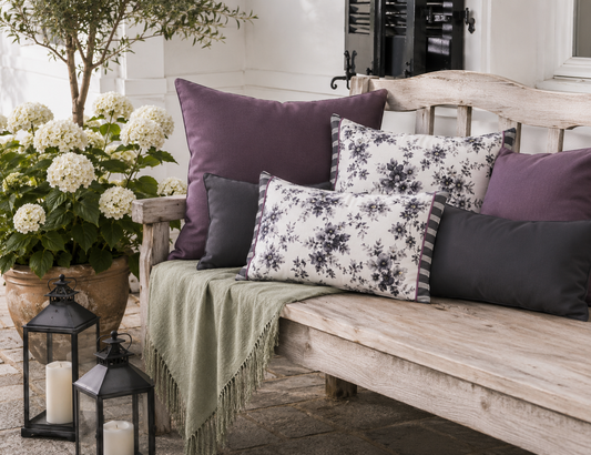 Dekokissen-Kombination in Aubergine, Anthrazit und Weiß auf einer rustikalen Gartenbank. Florale Kissen mit grauem Blütenmuster schaffen eine elegante Atmosphäre im Landhausstil auf Terrasse oder im Garten.