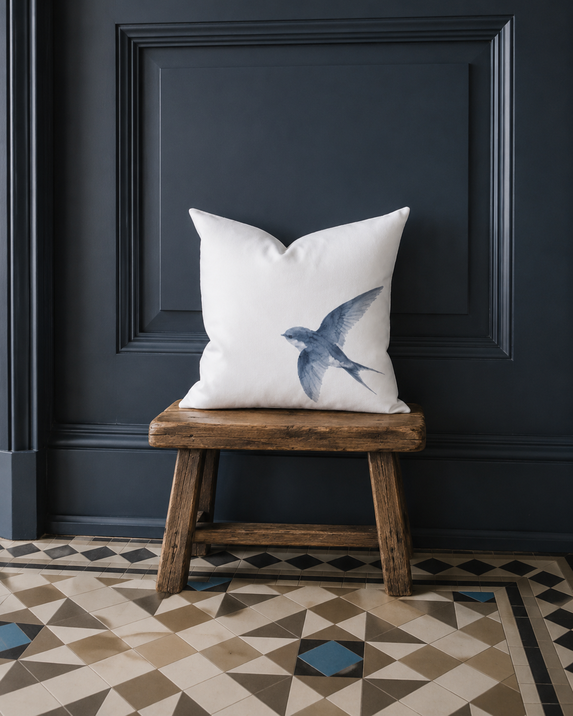Weiße Kissenhülle mit blauer Schwalbe als Aquarellmotiv auf einem rustikalen Holzhocker vor dunkelblauem Wandpanel im Landhausstil.