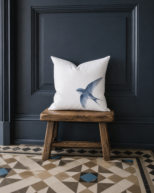 Weiße Kissenhülle mit blauer Schwalbe als Aquarellmotiv auf einem rustikalen Holzhocker vor dunkelblauem Wandpanel im Landhausstil.
