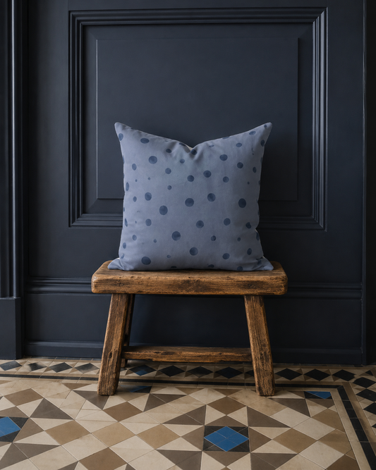 Kissenhülle Schwalbendots aus der Kollektion Schwalbe & Nebelblau auf einem rustikalen Holzhocker vor dunkelblauem Wandpanel im Landhausstil.